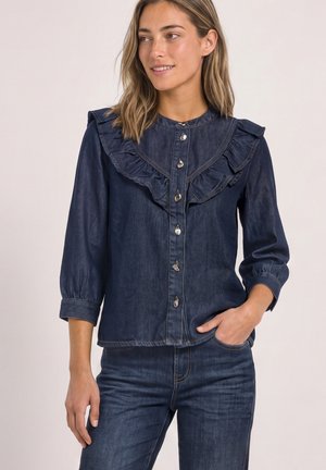 Femme portant une chemise en denim bleu foncé à col à volants et boutons argentés, assortie à un jean en denim assorti, souriante et regardant sur le côté.