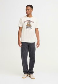 Hvit grafisk t-skjorte med en dovendyr i hettegenser og teksten "STØTT LOKALE GATEKATTAR", kombinert med mørke jeans og beige sneakers.