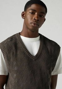 Gestrickte, ärmellose braune Strickweste mit Rautenmuster und V-Ausschnitt, übereinander getragen mit einem weißen Langarmshirt.