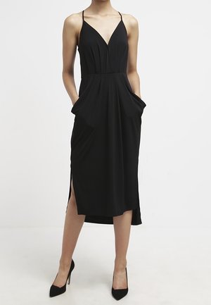 Mujer con un vestido midi negro sin mangas con tirantes finos, aberturas laterales, bolsillos y zapatos negros de tacón alto.