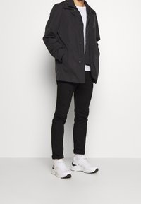 Czarna wodoodporna kurtka z klasycznym kołnierzem, zapinana z przodu na zatrzaski, noszona na białej koszulce, zestawiona z czarnymi spodniami i białymi trampkami.