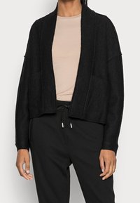 Czarny krótki kardigan wykonany z fakturowanej tkaniny, z otwartym przodem, długimi rękawami i dwoma przednimi kieszeniami, stylizowany na beżową bluzkę i czarne spodnie.