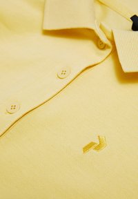 Polo giallo realizzato in tessuto texturizzato. Presenta un colletto, tre bottoni e un logo ricamato in un giallo leggermente più scuro.
