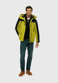 Green and black jacket with a hood, featuring two front pockets, full zip, and drawstrings. Paired with dark jeans and brown shoes.