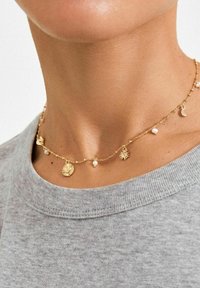Gouden ketting met bedels van een zon, maan en kleine parels, met een delicate ketting en getextureerde accenten, gedragen over een grijs shirt.