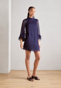 Navy blue dress with sheer long sleeves, ruffled cuffs, and a straight silhouette. Model wears black slide sandals and carries a small bag.