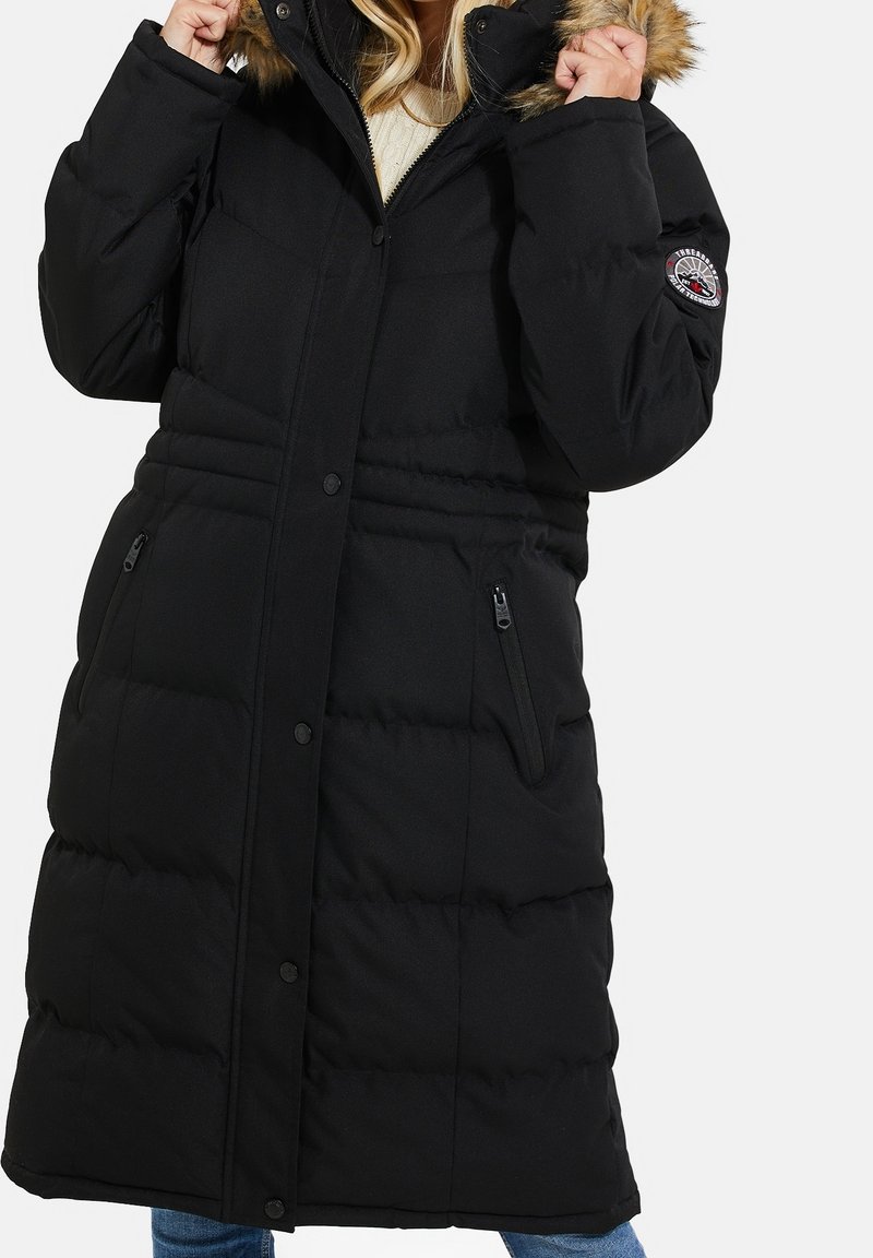 Femme ajustant la capuche d'un long manteau noir matelassé avec une bordure en fourrure sur la capuche et des poches latérales zippées.