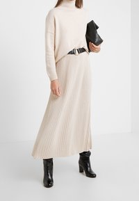 Pull en tricot côtelé beige à col haut, associé à une jupe maxi plissée crème, rehaussé d'une ceinture noire et de bottines noires à talons.