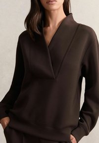 Suéter marrón de cuello en V profundo, con mangas largas y corte relajado. Hecho de una tela suave y lisa con un discreto detalle de costura.