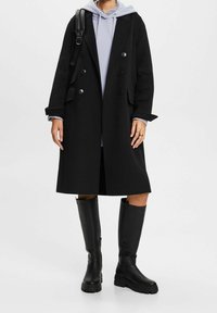 Abrigo negro de lana con mezcla de doble botonadura y grandes botones, combinado con una sudadera gris claro y botas de goma negras de altura hasta la rodilla.