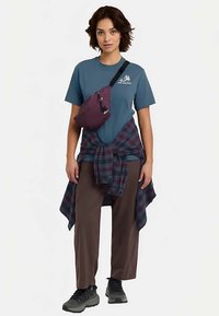 Maglietta blu con logo, pantaloni marroni, camicia a quadri legata in vita, borsa a tracolla viola e scarpe da ginnastica grigie. Outfit casual da esterno.