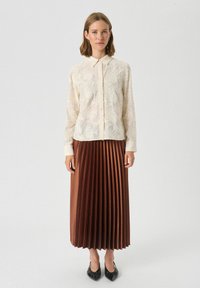 Lys beige broderet bluse med krave, sat sammen med en plisseret brun nederdel. Modellen har sorte spidse flade sko på, foran en ensfarvet baggrund.