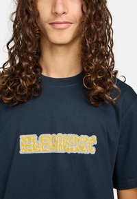 Mørkeblå bomulds t-shirt med et gult og hvidt abstrakt grafisk tekstdesign over brystet. Korte ærmer og rund hals.
