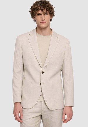Giovane uomo con capelli ricci che indossa un blazer beige chiaro sopra un maglione e pantaloni abbinati, in piedi davanti a uno sfondo semplice.