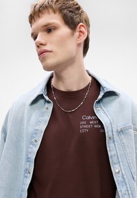 Chaqueta de mezclilla, de color azul claro, llevada sobre una camiseta marrón con texto blanco. Se puede ver un collar de tono plateado con eslabones rectangulares.