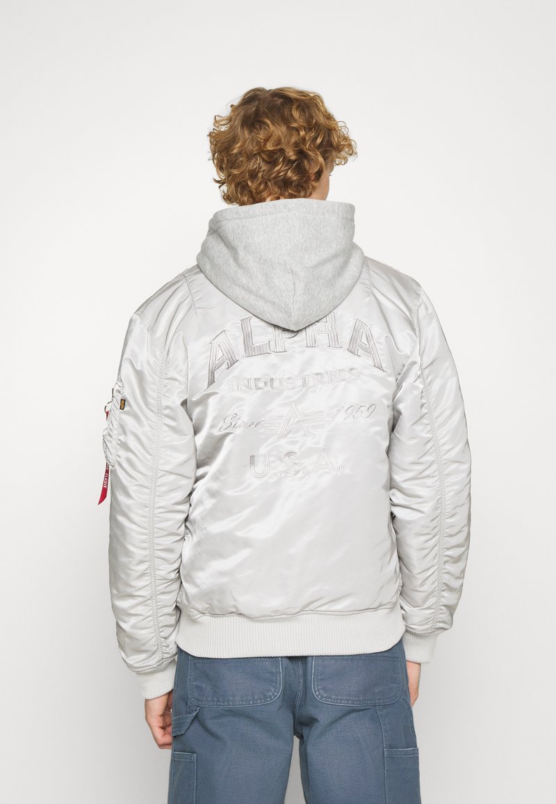 Blouson en satin argenté avec une texture matelassée, des poignets côtelés et une capuche grise. Présente un logo brodé et une étiquette rouge sur la manche.