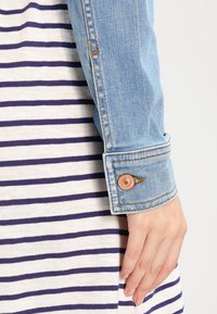 Denimtakki hihan messinkinarulla, kerrostettuna valkoisen ja tummansinisellä raidallisella kankaalla. Teksturoitu denim, räätälöity malli.