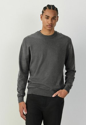 Mladý muž s vrkočmi, oblečený v tmavosivom dlhom rukáve svetri a čiernych nohaviciach, stojaci s jednou rukou v vrecku na svetlom pozadí.