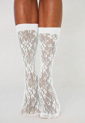 Jambes portant des chaussettes blanches en dentelle montantes avec des motifs floraux sur un fond gris uni.