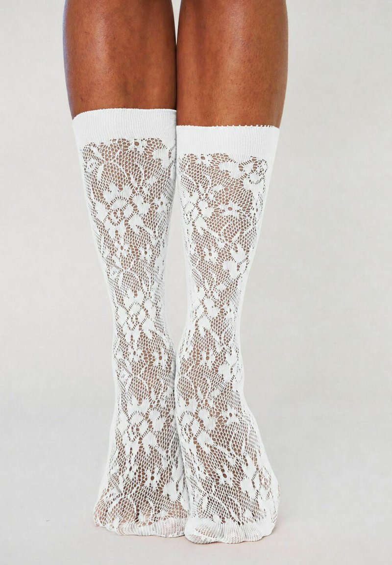 Jambes portant des chaussettes blanches en dentelle montantes avec des motifs floraux sur un fond gris uni.