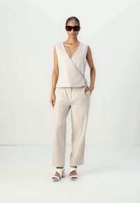 Woman wearing beige sleeveless wrap top, matching wide-leg pants, white heeled sandals, and oversized brown sunglasses, standing with one hand in pocket.