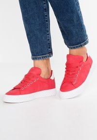 Pieds portant des baskets rouges avec des semelles blanches et des jeans bleus courts sur un fond blanc uni.