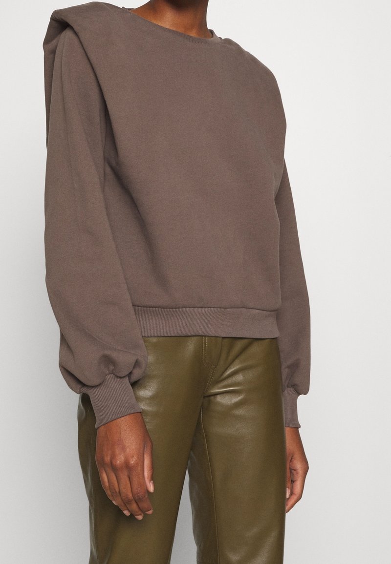 Personne portant un haut marron à manches longues avec des épaules bouffantes et un pantalon en cuir vert olive, debout devant un fond blanc uni.