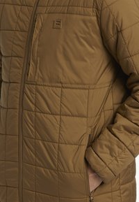 Brown quilted puffer jacket featuring a zippered front, textured fabric, side pockets, and a chest logo patch. Worn with hands in pockets.