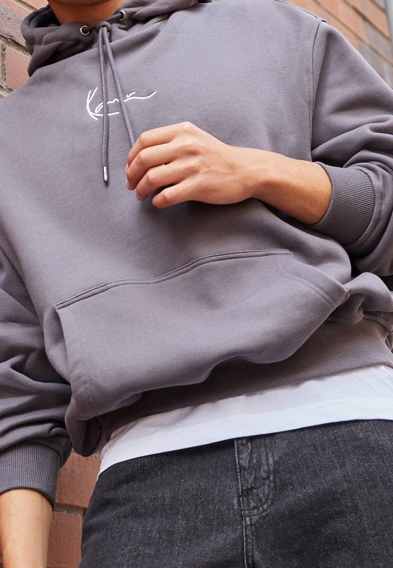 Personne portant un sweat à capuche gris avec une poche avant et un logo manuscrit blanc, reposant une main près de la poitrine, contre un mur en briques.