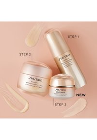 Tre Shiseido-hudvårdsprodukter: en krämburk, en hög serumflaska och en liten ögonkräm, alla i mjukt rosa och guld förpackningar, på en rosa bakgrund.