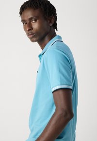 Νεαρός άνδρας με κοντά σγουρά μαλλιά, φορώντας ένα ανοιχτό μπλε πόλο μπλουζάκι με λευκή και μπλε ρίγα στις συνεχίσεις του γιακά και τα μανίκια, κοιτάζοντας πλάγια.