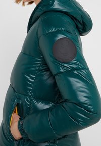Femme portant une veste matelassée vert foncé brillante avec capuche, main dans la poche, et un patch noir rond sur le haut de la manche.