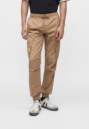 Mann trägt beige Cargohosen mit elastischen Bündchen und schwarz-weißen Sneakers, steht vor einem weißen einfarbigen Hintergrund.