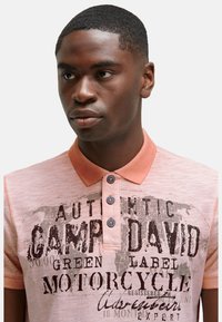 Licht koraalkleurig poloshirt met een zachte textuur, voorzien van een drie-knopssluiting, contrasterende kraag en grafische tekstprint in donkerbruin.