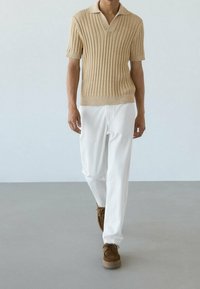 Polo à manches courtes en tricot côtelé beige, associé à un pantalon blanc et des chaussures à lacets marron, sur fond minimal.
