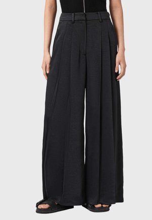 Musta, laskostetut, leveälahkeiset housut, jotka on valmistettu teksturoidusta kankaasta. Niissä on korkea vyötärö ja sileä etuosa, yhdistettynä mustiin avojalkaisiiin sandaaleihin.