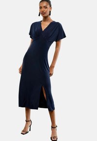 Navyblaues, knielanges Kleid mit kurzen Ärmeln, einem V-Ausschnitt und einem Seitenschlitz. Hergestellt aus glattem Stoff mit gerafften Details an der Brust.