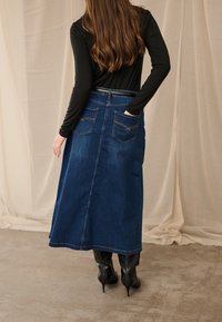 Moteris stovi, viena ranka įkišusi į užpakalinę kišenę, dėvi ilgą mėlyną džinsinę sijoną, juodą ilgomis rankovėmis palaidinę ir juodus kulnuotus batus, stovi ant smėlio spalvos grindų.