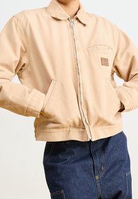 Giacca beige in cotone con colletto, chiusura con zip, tasche anteriori e una piccola toppa marrone con logo sul petto. Tessuto strutturato, vestibilità casual.