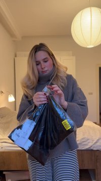 Γυναίκα με γκρι πουλόβερ και ριγέ παντελόνι ανοίγει ένα μαύρο πλαστικό πακέτο σε ένα υπνοδωμάτιο με απαλό φωτισμό, με κρεβάτι και στρογγυλό φωτιστικό.