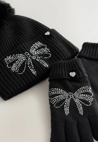 Black knit hat and gloves set with silver rhinestone bow designs. Hat features a pom-pom; gloves have ribbed cuffs and heart-shaped accents.