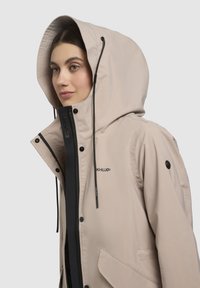 Jachetă impermeabilă color beige cu o glugă mare, fermoar negru contrastant, nasturi tip capse și buzunare laterale; are o textură netedă și cordoane ajustabile.
