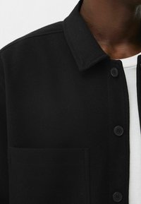 Czarna wełniana kurtka z kołnierzem, przednią kieszenią i czarnymi guzikami. Tkanina ma gładką fakturę i dopasowany krój.