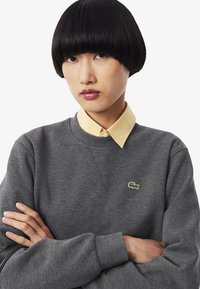 Giovane con un taglio di capelli bob nero liscio indossa un maglione grigio Lacoste sopra una camicia gialla con colletto, braccia incrociate, espressione neutra.