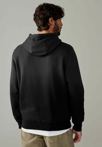 Black hoodie with a kangaroo pocket and adjustable hood, made of soft fabric. Ribbed cuffs and hem add texture. Worn over a white shirt.