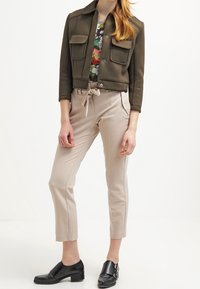 Olivengrøn cropped jakke med to brystlommer, parret med beige tapered bukser og sorte spænde sko. Blomsterprint top synlig.