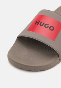 Grijze slide-sandaal met een getextureerde band voorzien van een rode rechthoek en het zwarte "HUGO"-logo. Zachte, gladde binnenzool met een lichte gripstructuur.