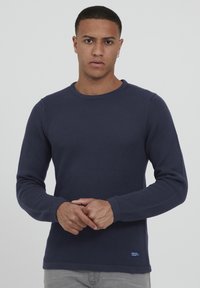 Pulover texturat de culoare bleumarin, cu manșoni lungi, guler rotund și manșete cu nervuri. Prezintă un mic patch cu logo pe partea inferioară.