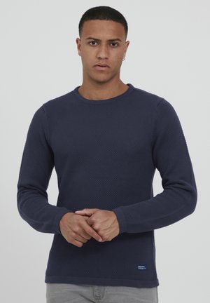 Pull marin en bleu marine avec texture, à manches longues, col rond et poignets côtelés. Présente un petit patch avec logo sur le côté inférieur.