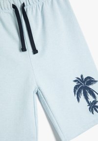 Lysblå bomuldsshorts med en sort snøre og et palme-træ grafik trykt i marineblå på venstre ben. Blød tekstur og afslappet design.